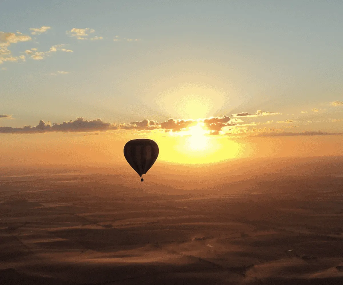 A silhouette of people in a hot air balloon, looking out at a sunrise and a hot air balloon in the distance