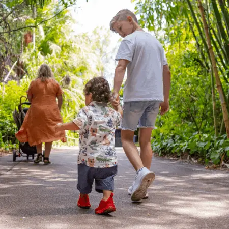 A toddler, older sibling holding hands, mum walking in front with a pram. Lush green surroundings and a concrete path